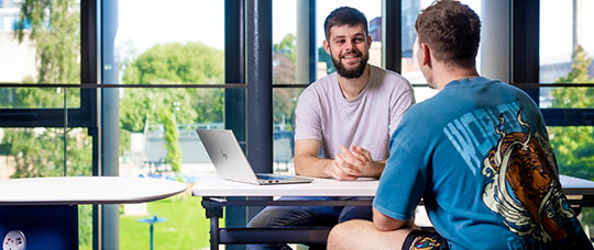 Two students sitting at a table with one laptop smiling.