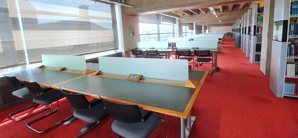 Quiet study spaces in the Library, rows of 8 seater tables with windows and stacks of book stock.