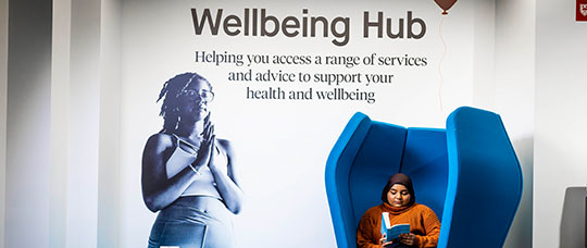 Student sitting in large blue chair reading a book with a wall sticker behind the chair with the text "Wellbeing Hub"
