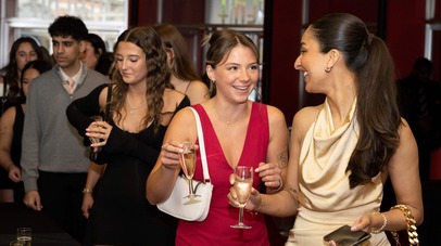 2 female students in right foreground, dressed in smart dresses and smiling at each other, with other students in the background.