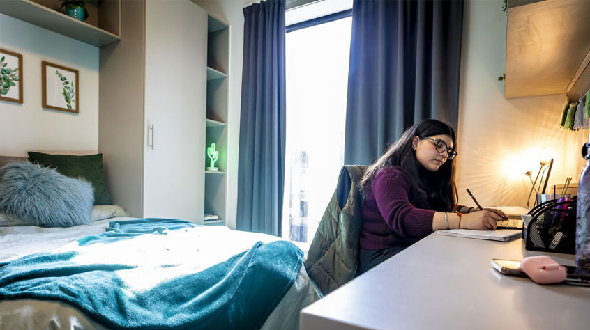 A student studies at her desk in her room. Light streams in through window