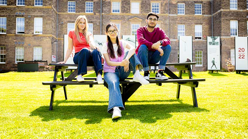 Photo of three students, two female, one male, sat on a picnic bench looking towards camera
