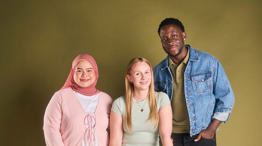 Three prospective students standing together in a studio against an olive-green background. One person wears a pink headscarf and cardigan, another wears a light green top, and a third wears a denim jacket over a green shirt. The group faces the camera in a welcoming recruitment-style portrait.