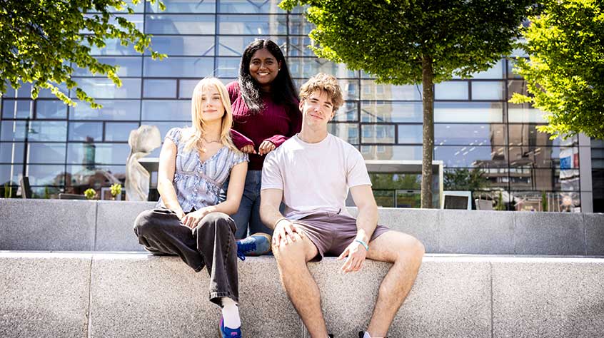 Three people sit together on a low concrete wall outdoors in front of a modern glass building. One person in a light outfit sits on the left, another in a white T-shirt and shorts sits on the right, and a third person in a darker top stands slightly behind them with hands resting on their shoulders. Green trees frame the scene, and bright sunlight casts clear shadows.