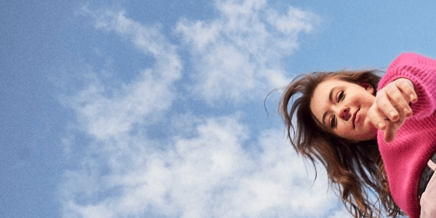 A young woman looks down at the camera while she strides through campus, with a bright blue sky and tall building as a backdrop