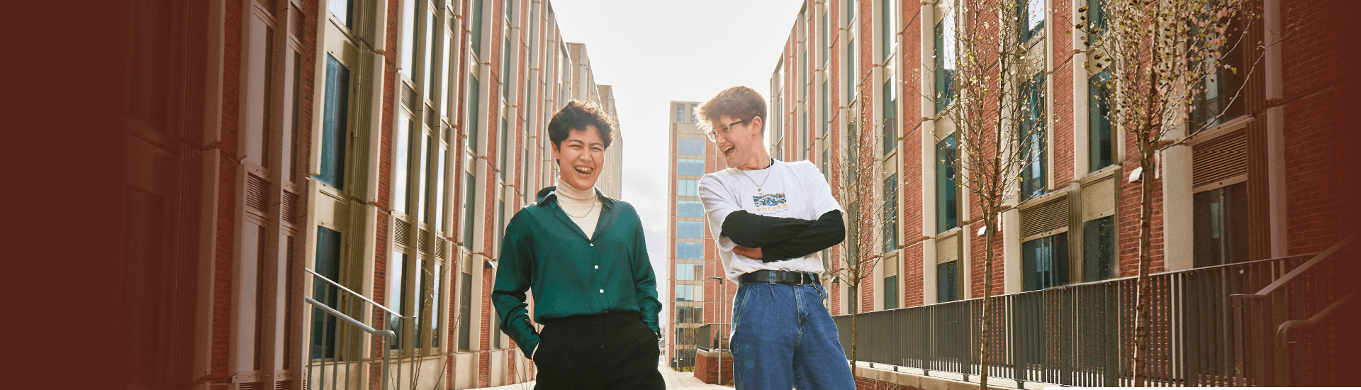 Two young people stand between tall buildings. One has hands in pockets the other has arms folded, both in fits of giggles