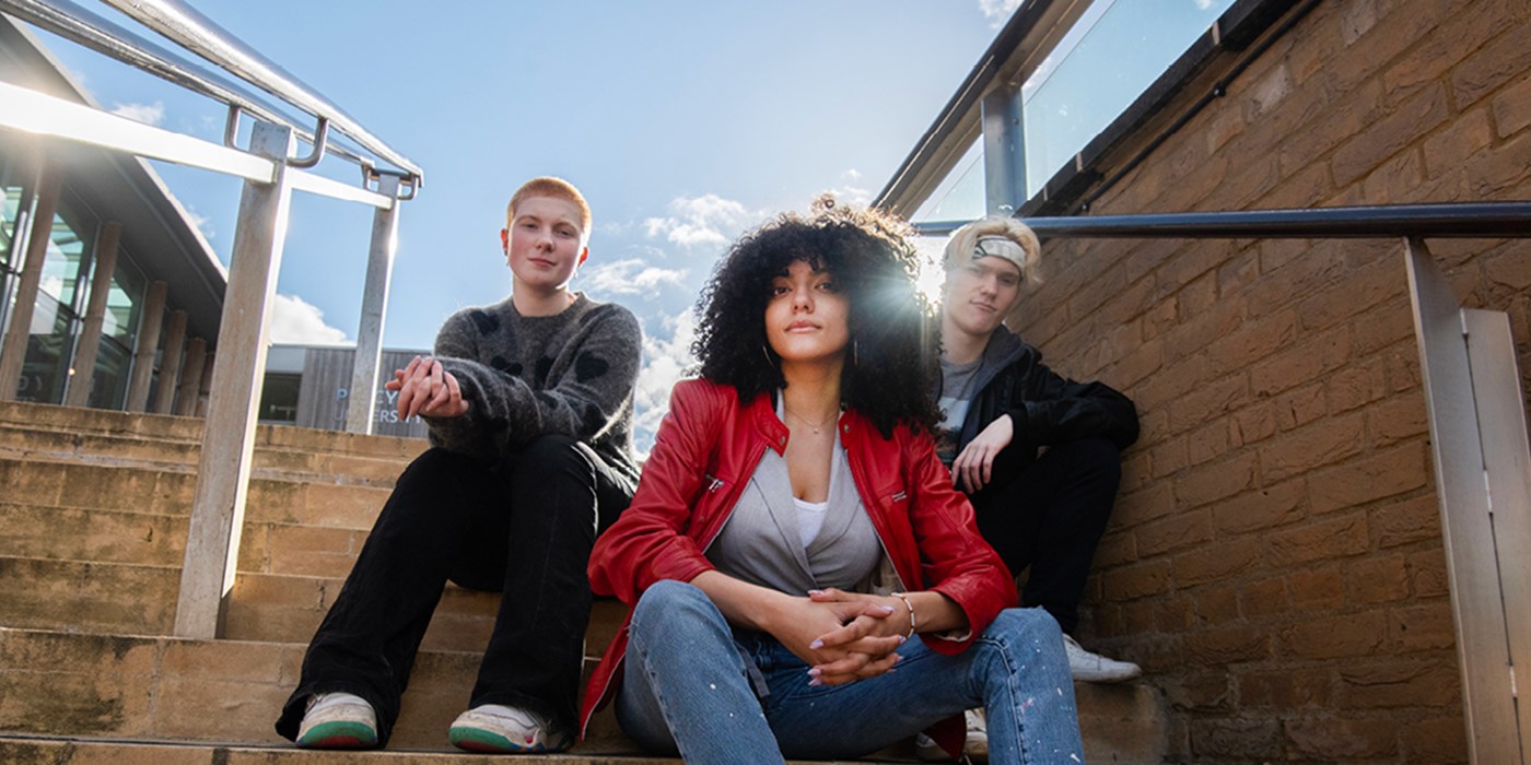 Three young people look down at camera while sitting on steps with buildings, bright blue sky and sunlight as backdrop