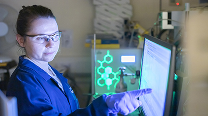 A scientist points at a computer screen with a bright green hexagonal diagram on another screen in the background