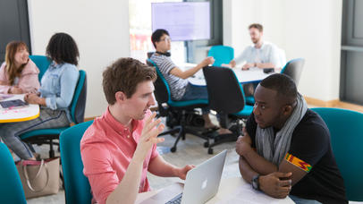 Image of student academics working and conversing together at desks with laptops and notebooks