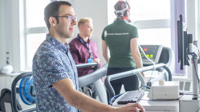 Photo of an academic at a computer while a subject is using a treadmill, with instruments reading his vitals and another academic checks the machinery output