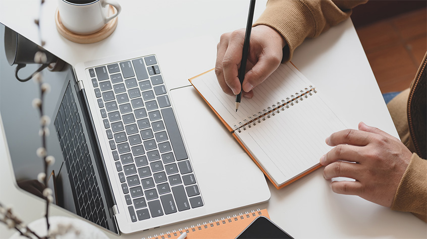 Top down view of a person sat at a table writing in a notebook with their laptop open and a mug with drink almost out of shot.
