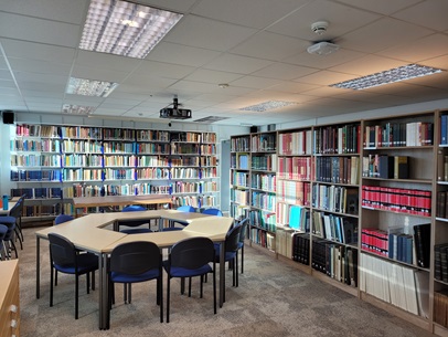 Photo of seminar space in a library with bookshelves on the right and in the background. In the foreground is a small group of desks and chairs arranged in a circle for a seminar. 