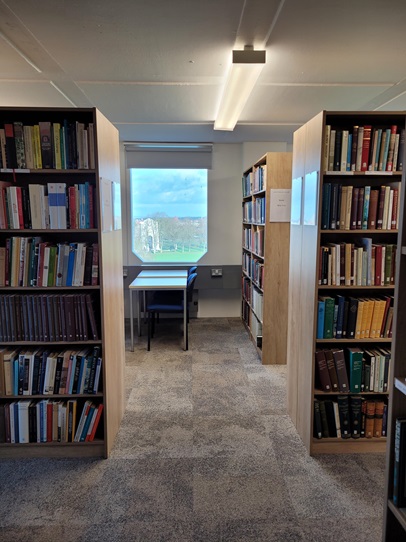 Photo of book shelves in a library, with an empty desk and chair centred in the image below a window. Through the window, the cenotaph in Victoria Park, Leicester is visible. 