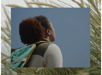 A person with curly hair and a teal backpack gazes upward beneath a clear blue sky, framed by sunlit grasses; the scene feels calm and hopeful.