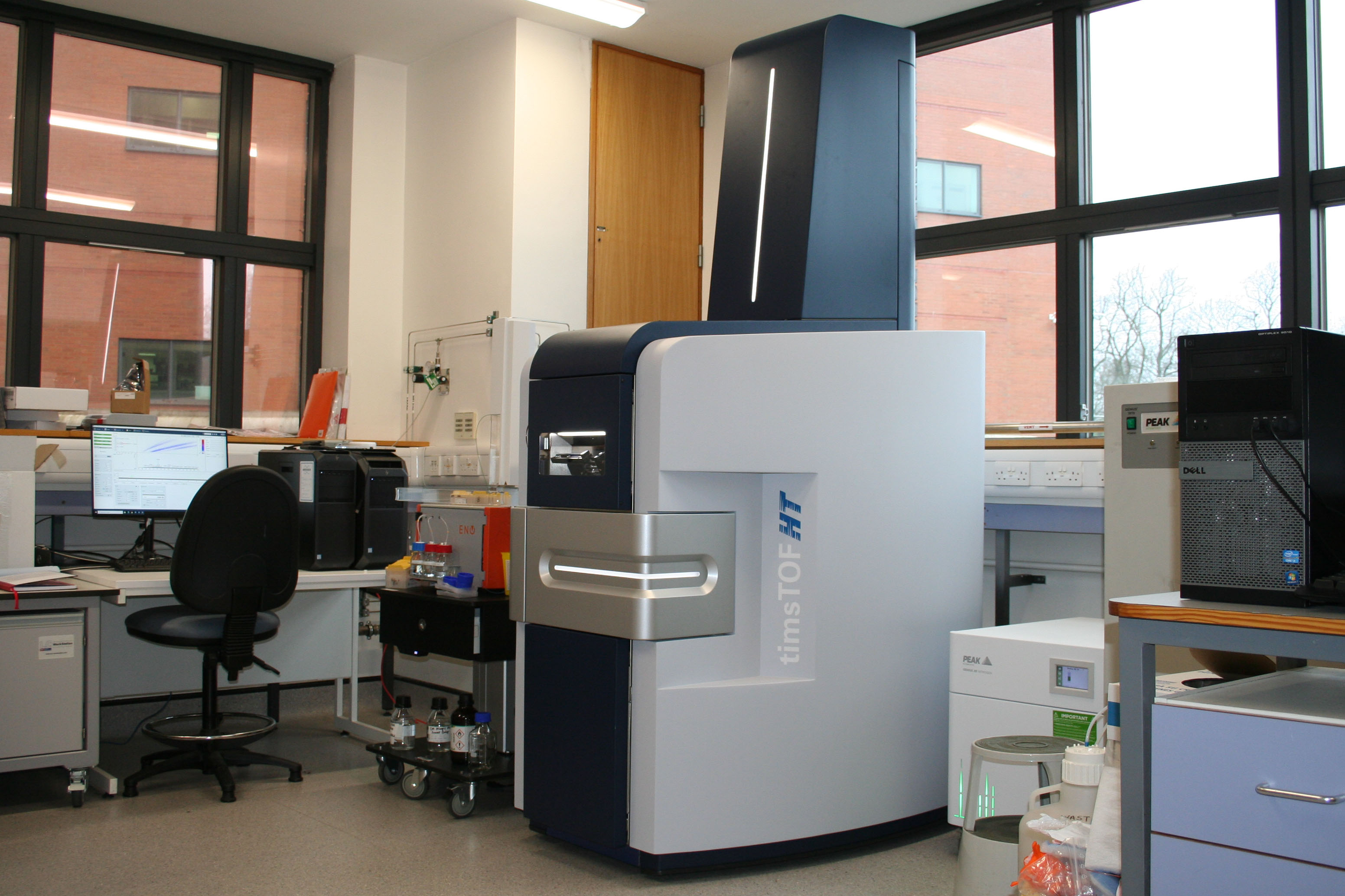 Laboratory with a large, modern mass spectrometer labeled "timsTOF HT." Nearby desk with computer, chair, equipment. Bright, organized space.