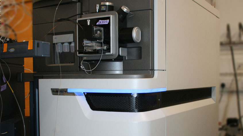 A large, modern mass spectrometer in a lab setting, featuring a sleek, grey design with visible wiring and components, emits a soft blue glow.