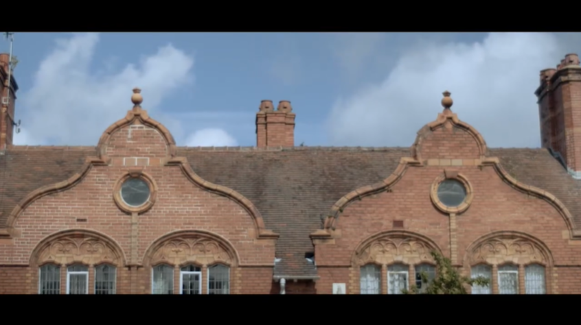 The facade of a red brick building with tiled roof and a chimney. Blue sky with clouds in the background