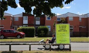 A man rides along a residential street on a tricycle with a bright yellow banner attached that reads 'The Campaign for Empathy Celebrates Arbury Carnival' Part of the art project Campaign for Empathy: North Cambridge, Open House by artist Enni-Kukka Tuomala