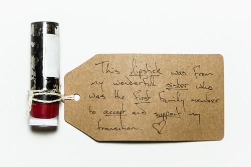 Red lipstick (closed, with lid on). A brown museum tag is attached to it. The tag reads: "This lipstick was from my wonderful sister who was the first family member to accept and support my transition". There is a small heart doodle in the corner.
