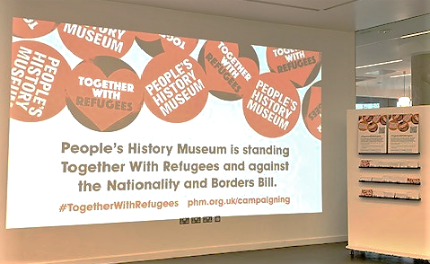 Image of a slide deck projected onto a white wall. The slide says 'People's History Museum is standing together with refugees and against the Nationality and Border Bill'. Orange museum logo stickers and stickers saying 'together with refugees' can be seen above the slide title.