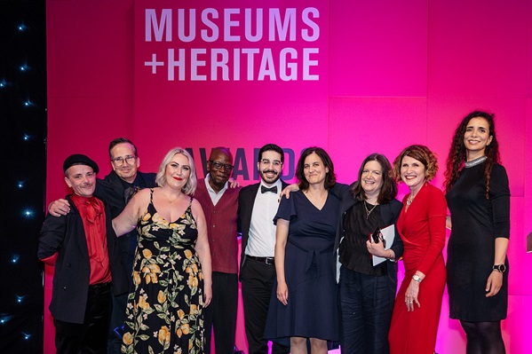 Nine people posing for a photo in front of a red backdrop with the Museums + Heritage Awards Logo