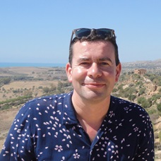 A headshot of Professor Gowan Dawson, a man, smiling at the camera. He has sunglasses on his head and a dark blue shirt on, and in the background is a sunny landscape dotted with old buildings.