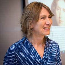 A headshot of Dr Julian North. She is smiling at something to the right of the camera, she has dark blonde hair which is tied up and is wearing a blue dotty shirt. Behind her is a screen with an image of George Eliot. 