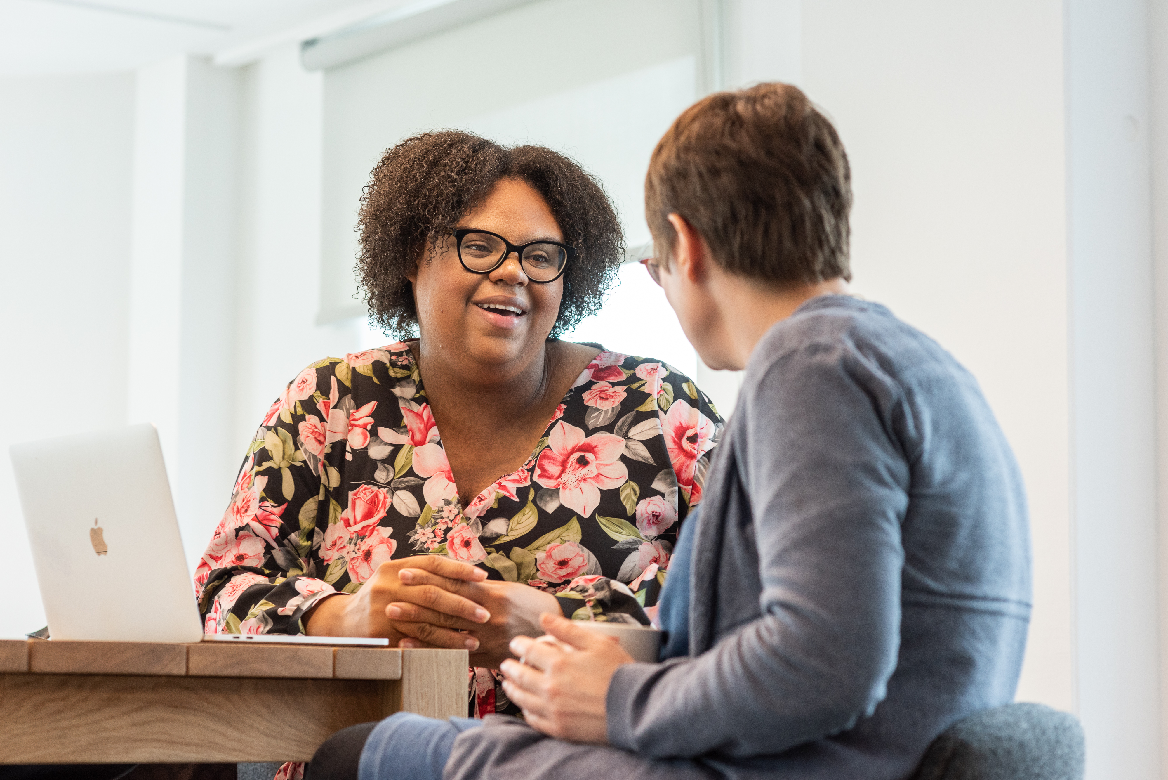 Two women are sitting at a high table and chatting. One is facing towards the camera and is a smiling young black woman, the other, a white woman, is facing away from the camera and is holding a mug. There is an open laptop on the table in front of them.  