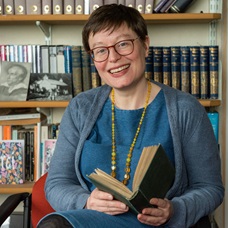 A headshot of Dr Felicity James. She is sat down holding a book and their is a full bookcase behind her. She has dark brown short hair, is wearing a blue dress and cardigan, and is smiling. 