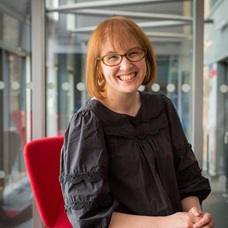 A headshot of Dr Claire Wood. She has a strawberry blonde bob and is wearing a black dress. Claire is leaning on a table with a red chair and a modern window behind her. 