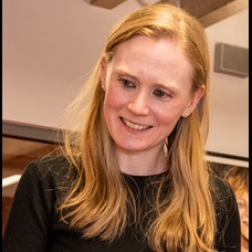 A photograph of Claire Brock, a white woman with strawberry blonde hair and who is wearing a black top. She is shown smiling at something off camera to the left of her. 