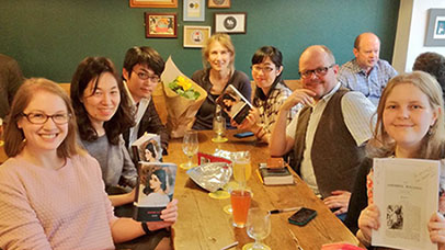 Seven people are sat round a table smiling at the camera, a couple of them are holding copies of the book Romola as they have been reading together. 