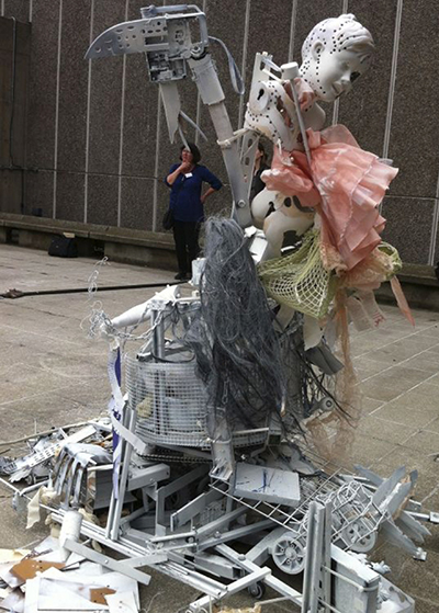 An sculpture made of destroyed personal belongings; a tall structure of plastic, metal, wire, and clothing topped by a large plastic axe and a mannequin head dotted with holes
