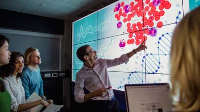 Man presenting in front of a large screen with data on it.