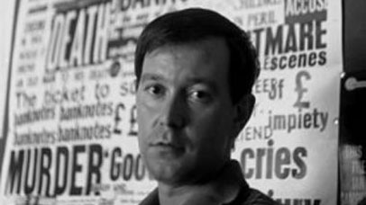 Joe Orton looking down the camera in front of a poster for Loot.