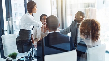 two business people shaking hands over a deal with other members of the team sat in an office