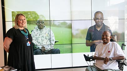 Two people are in front of a large screen, one is standing, one is sat in a wheelchair. There are two people on screen, one person is the same wheelchair user in front of the screen, on screen they are in a field, the other person on screen is stood in the same field.