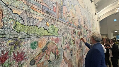 An angled perspective of piece of wall art, from ceiling to floor and covering the entire width of the wall. In black, printed outlines, it depicts the sky with birds, a city scape below, and the ground with coyotes jumping in amongst the bushes while surrounded by coyote bones. The image has been ‘coloured in’ using crayoned shaped like coyote bones, with many colours overlaying each other, with different additions by a number of contributors. The higher portions are less colourful, as they are not reachable without a ladder or similar apparatus. People are stood in front of the wall, to the right of the image, adding their own contributions, with JJ Hunt, a man with grey hair and beard, dressed in a royal blue suit jacket and black trousers, in the foreground colouring in the wall with a coyote bone-shaped crayon.