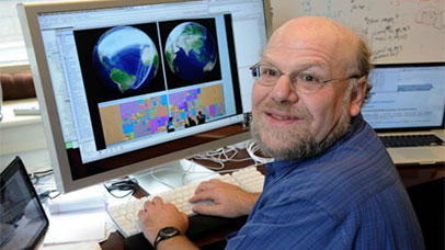 James Hendler is smiling and facing the camera whilst sitting at a desk on the computer, on the screen there are two images of planet earth, below these images are an array of colourful boxes with silhouettes of people in the foreground 