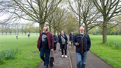 Photograph of 5 people walking towards the camera down a tree-lined path, with a green park landscape either side. The walkers are all wearing headphones as they take part in the events immersive sound walk. 
