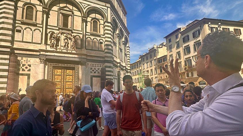 Man with students communicating with his hands stood in front of a building in a busy tourist area in a european town / city.