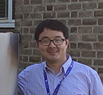 Dr Jiamiao Hu standing next to a UoL sign, wearing a blue shirt, beige trousers and black shoes.