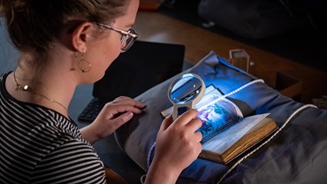 Woman reading an old book in a dark room with a magnifying glass and a light