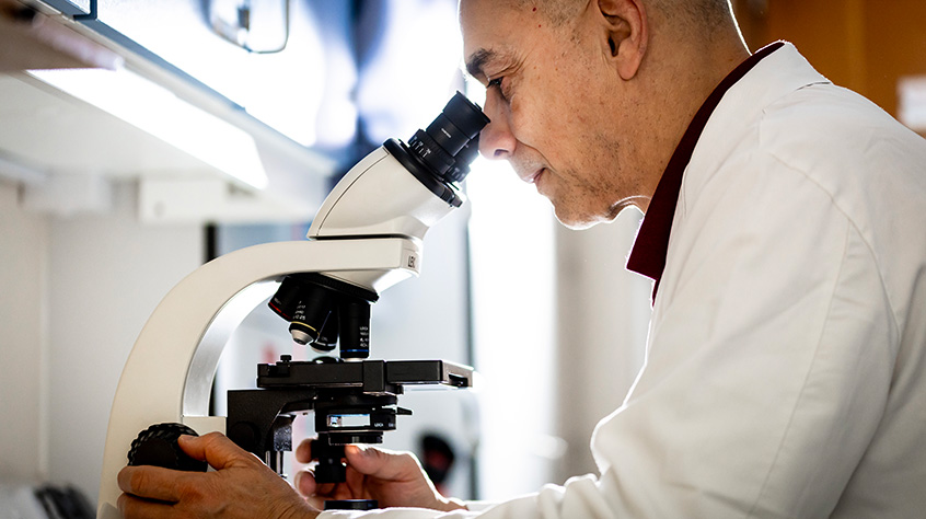Man in a  lab coat looking through the lens of a microscope.