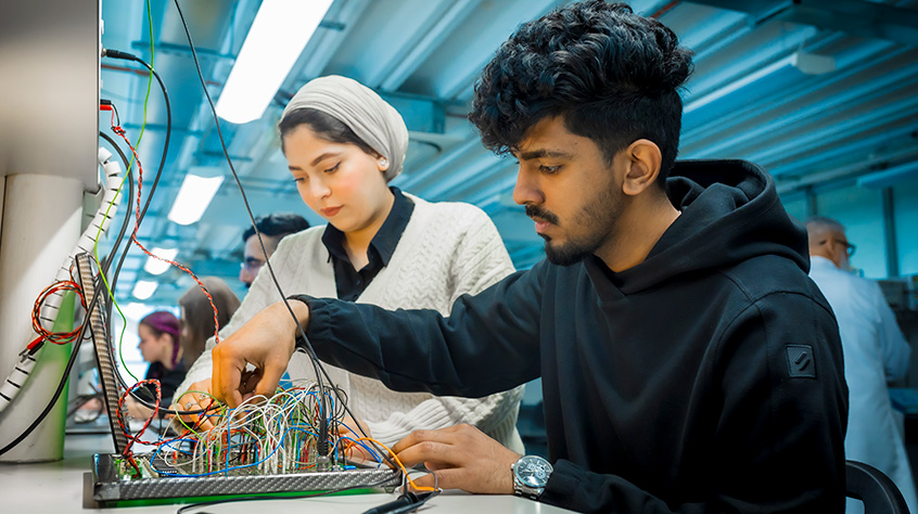 Two students, male and female sat at table in a workshop working on electronics