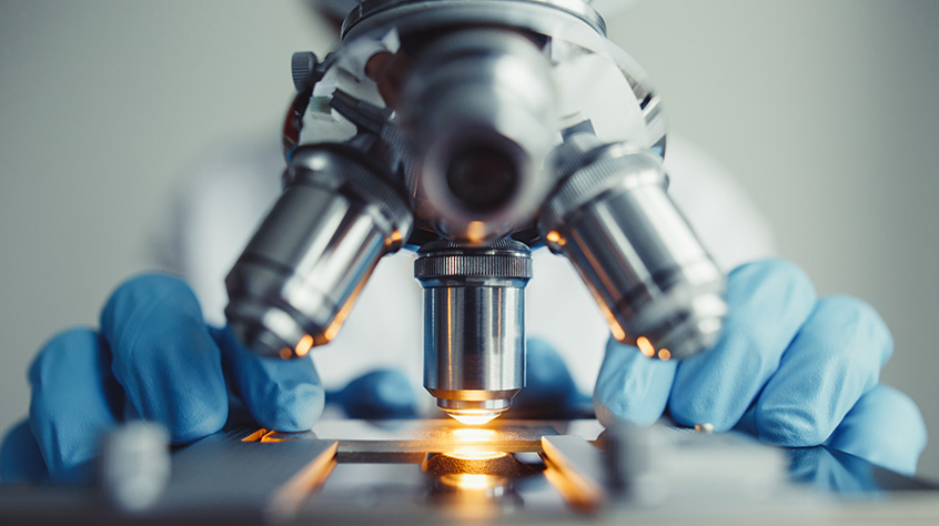 Person in labcoat adnw earing gloves is adjusting scientific equipment such as a microscope