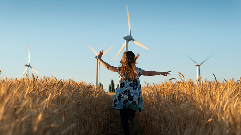 Person walking or dancing through a large field of crops, wind turbines can be seen beyond in the background.