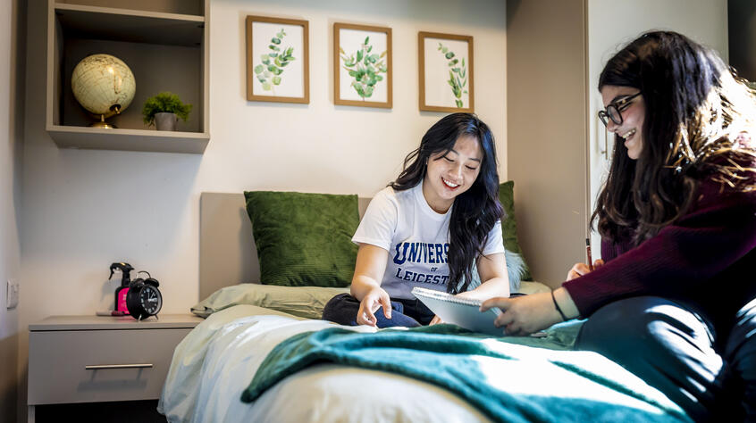 Two students sitting on a bed in their student accommodation, smiling and looking relaxed while reading