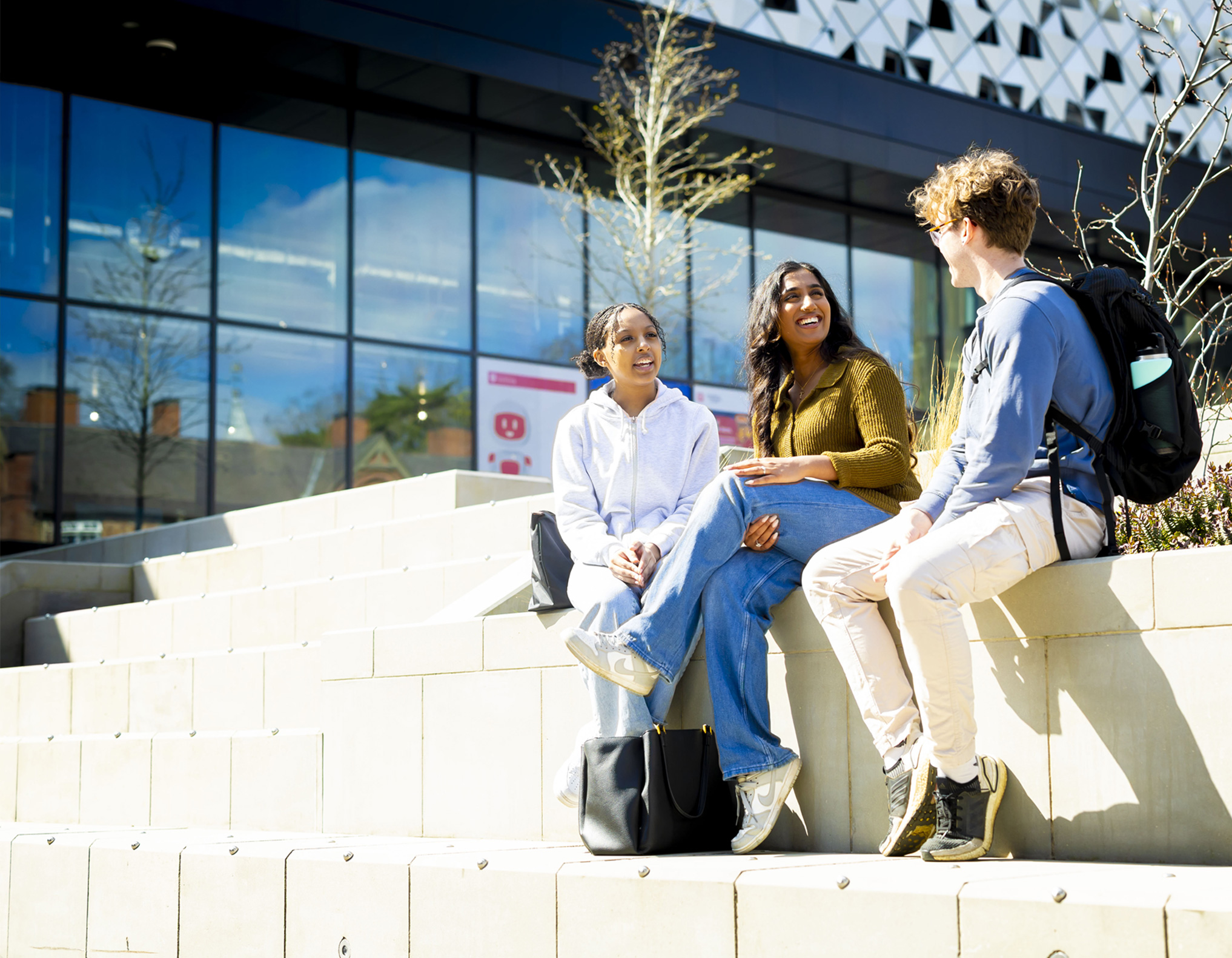 3 students smiling and chatting, sitting in the sunshine, outside Freemen's Bar & Kitchen in the Student Village