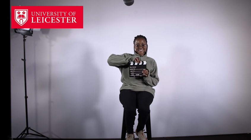 Student sitting in a studio and holding a clapperboard and smiling into the camera.